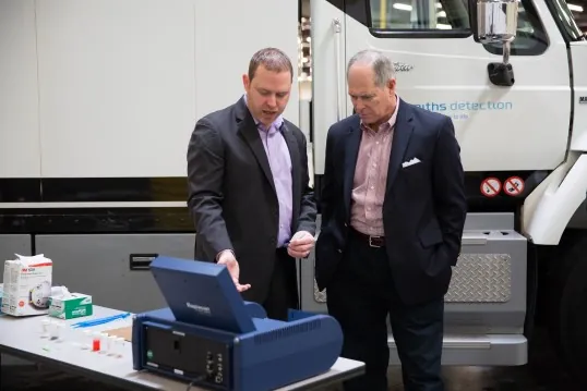 two men look at a research device, with a truck in the background.